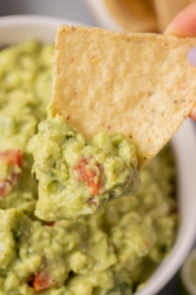 A tortilla chip taking a scoop out of a white bowl of guacamole