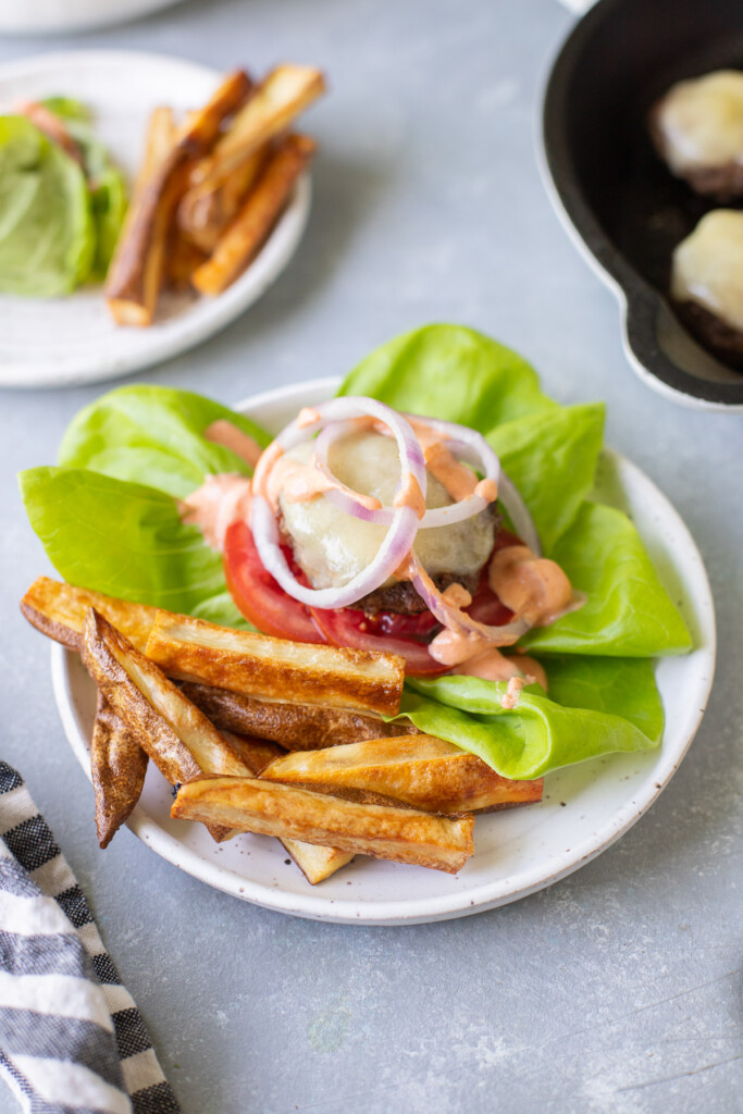 A close up of a lettuce wrapped burger on a white plate with fries off to the side