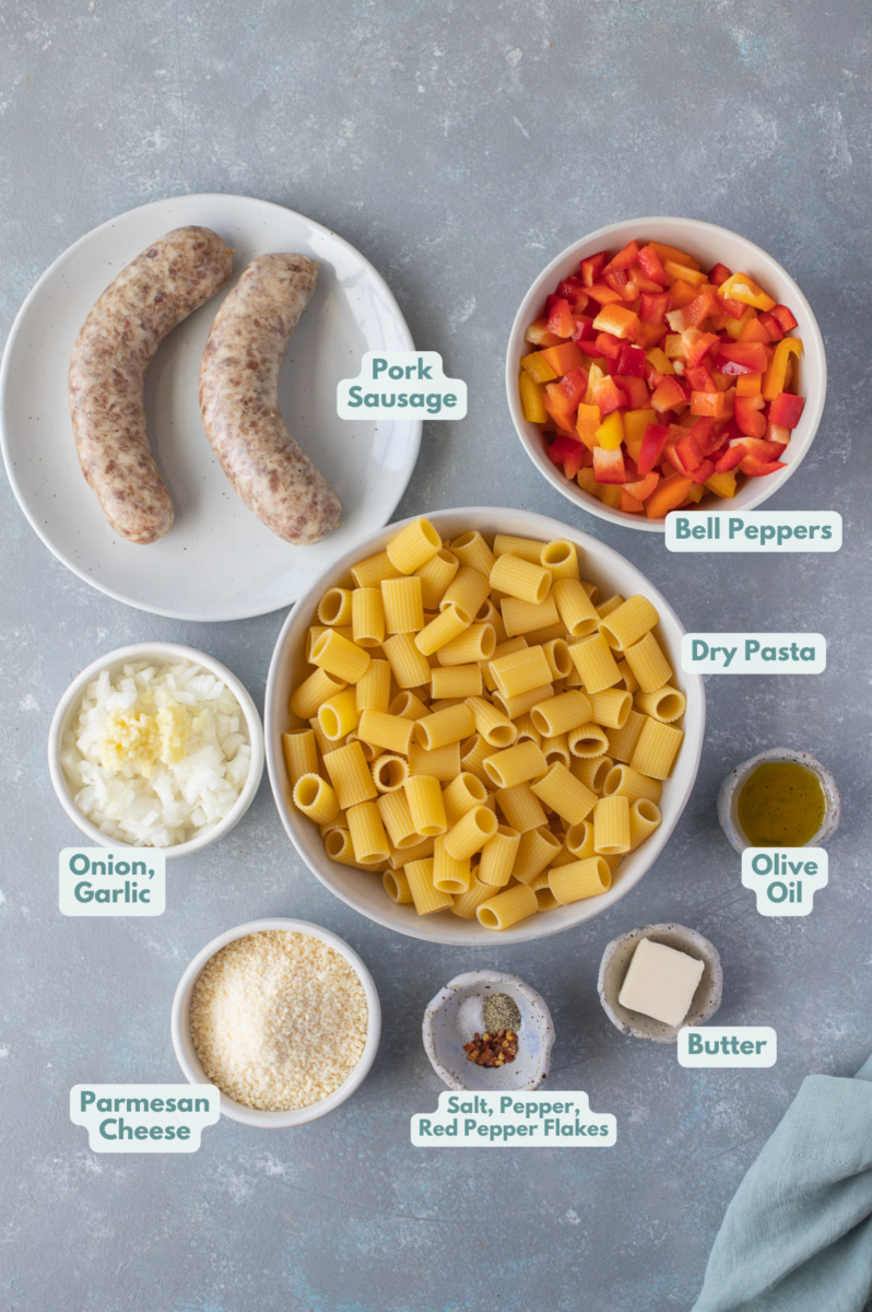 Ingredients for sausage and peppers pasta in white bowls on a grey background: ground sausage, olive oil, butter, onion, garlic, red pepper, salt, pepper, red bell peppers, dry pasta, parmesan cheese