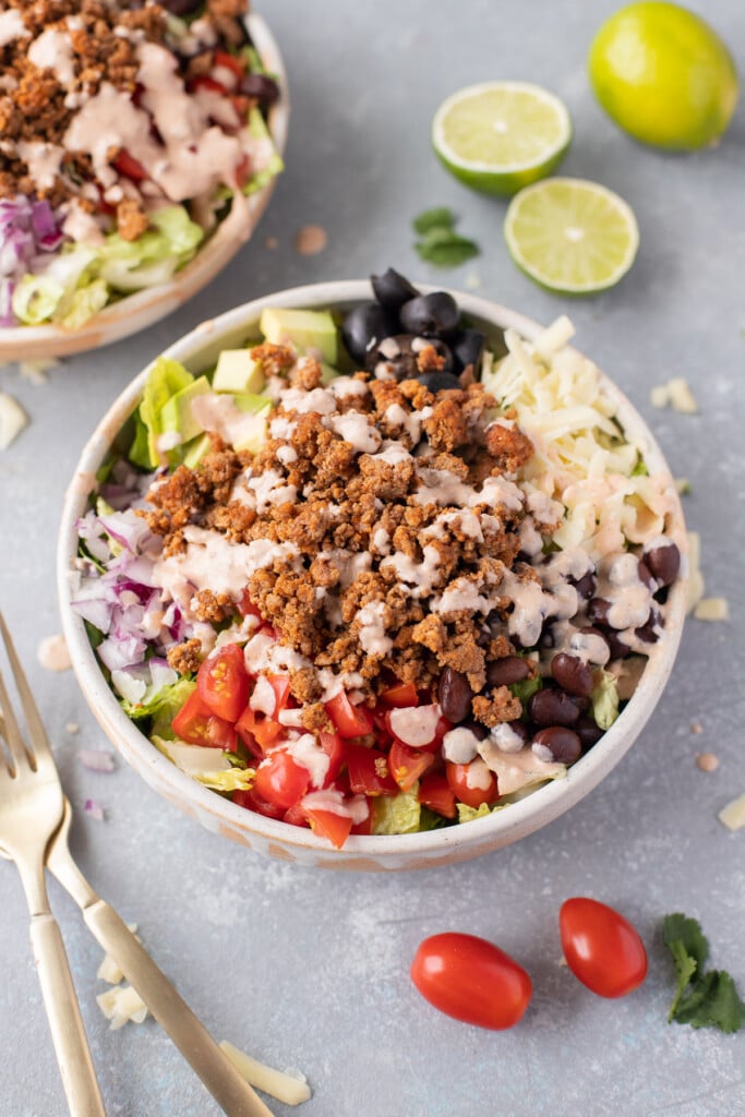A bowl of taco salad topped with taco dressing with toppings scattered around on a grey board