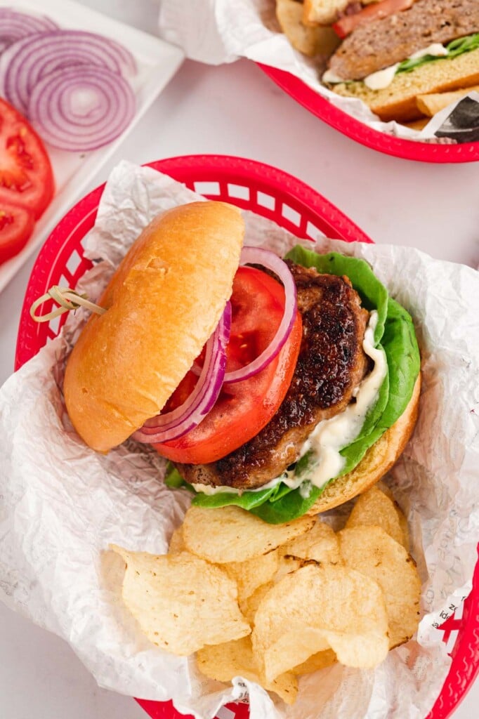 A turkey burger on a bun with tomato, red onion, lettuce and mayo in a red basket with a paper liner with chips