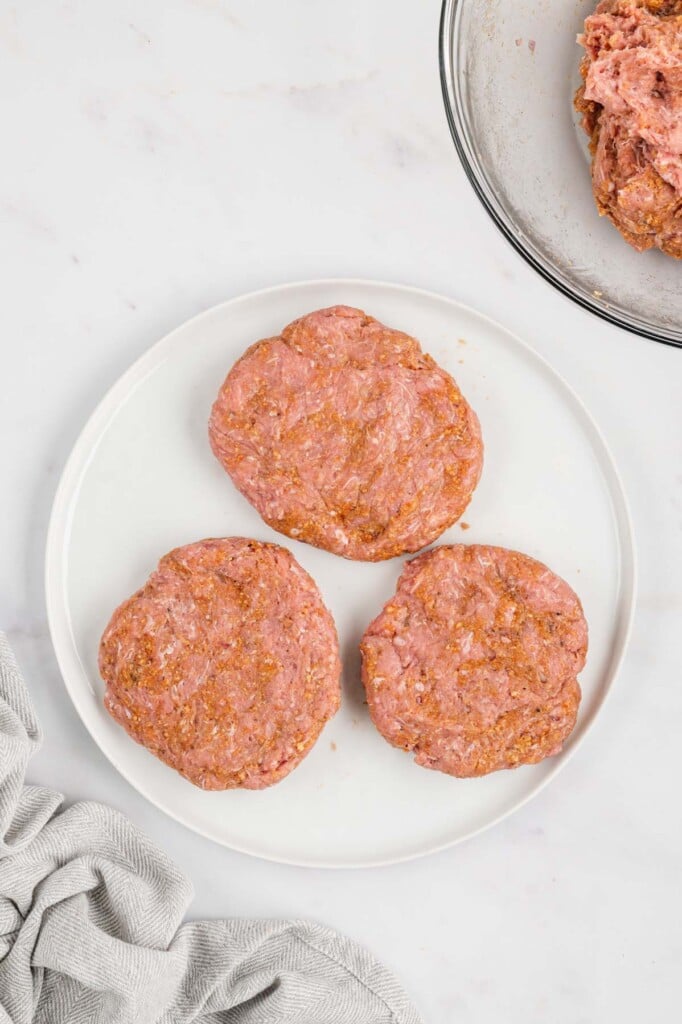 Three turkey burger patties on a white plate with a clear bowl of more meat on the side on a white background