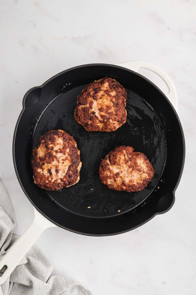 A cast iron pan with three browning turkey burgers in it on a white background