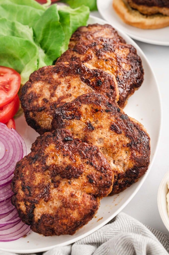 4 turkey burgers on a white platter with sliced red onions, tomatoes, and lettuce on the side