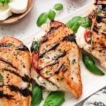 Three chicken caprese breasts on a white platter topped with balsamic glaze and fresh basil leaves with mozzarella and tomatoes on the side