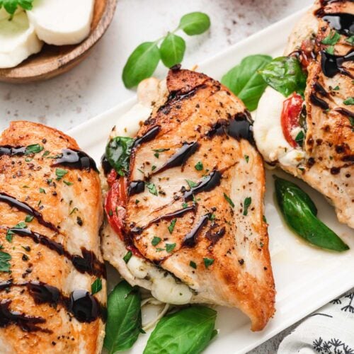 Three chicken caprese breasts on a white platter topped with balsamic glaze and fresh basil leaves with mozzarella and tomatoes on the side