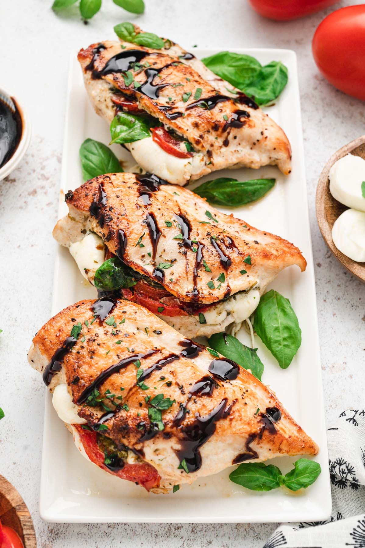 Mozzarella stuffed chicken breast on a white platter topped with balsamic glaze and fresh basil leaves with fresh mozzarella slices and tomatoes on the side.