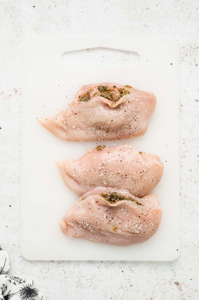 Three chicken breasts on a cutting board, stuffed with basil pesto and seasoned with salt and pepper