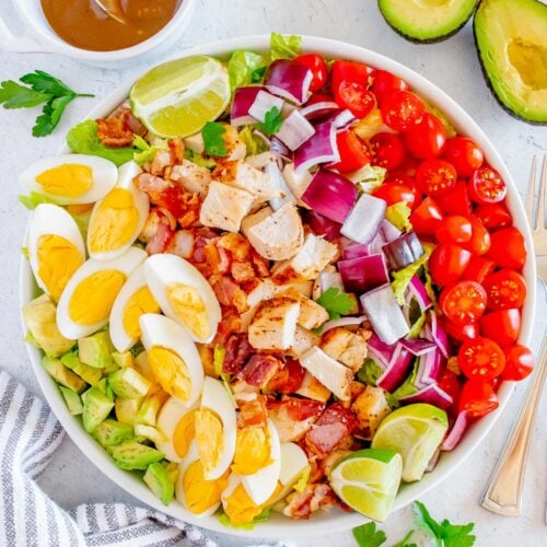 A chicken cobb salad with all the toppings in a white bowl with the Creamy Balsamic Salad Dressing in a small bowl and an avocado on the side