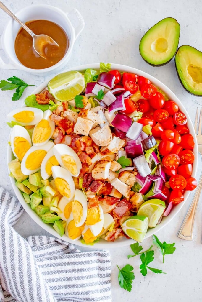 A chicken cobb salad with all the toppings in a white bowl with the Creamy Balsamic Salad Dressing in a small bowl and an avocado on the side