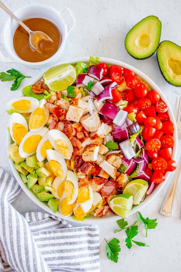 A chicken cobb salad with all the toppings in a white bowl with the Creamy Balsamic Salad Dressing in a small bowl and an avocado on the side