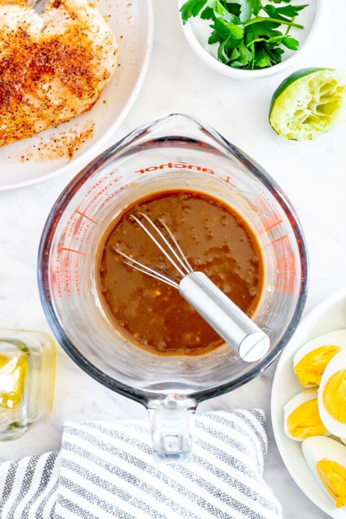 Creamy balsamic salad dressing in a clear pyrex measuring cup after being whisked with a whisk in the cup and ingredients for a chicken cobb salad surrounding them