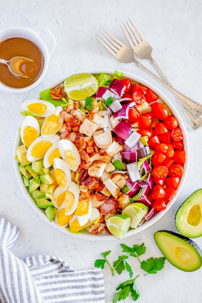 A chicken cobb salad with all the toppings in a white bowl with the Creamy Balsamic Salad Dressing in a small bowl, forks, and an avocado on the side