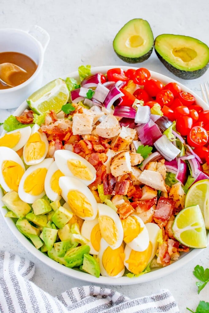 A Chicken cobb salad topped with red onion, cherry tomatoes, bacon and hard boiled eggs with a sliced avocado and creamy balsamic dressing in the background