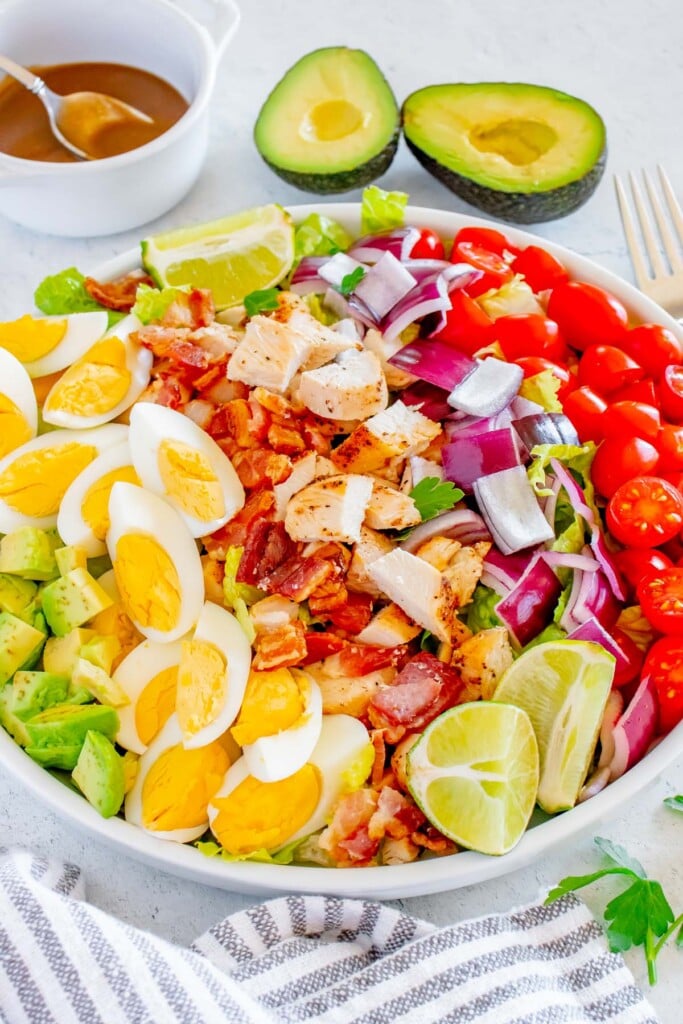 A Chicken cobb salad topped with red onion, cherry tomatoes, bacon and hard boiled eggs with a sliced avocado and creamy balsamic dressing in the background