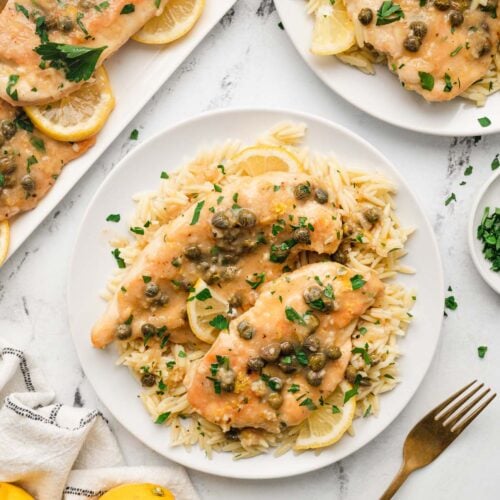 An overhead shot of chicken piccata on a white plate with a fork, lemons, another plate and platter on the side