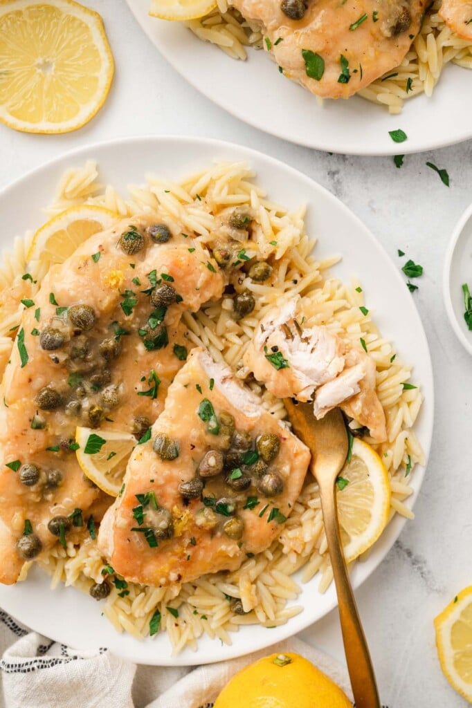An overhead close up of chicken piccata on a bed of orzo on a white plate with a fork taking a bite out of it with lemon slices on the side on a white background
