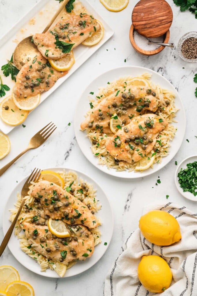 An overhead shot of two plates of chicken piccata with the platter of chicken on the side surrounded by lemons and spices on a white background