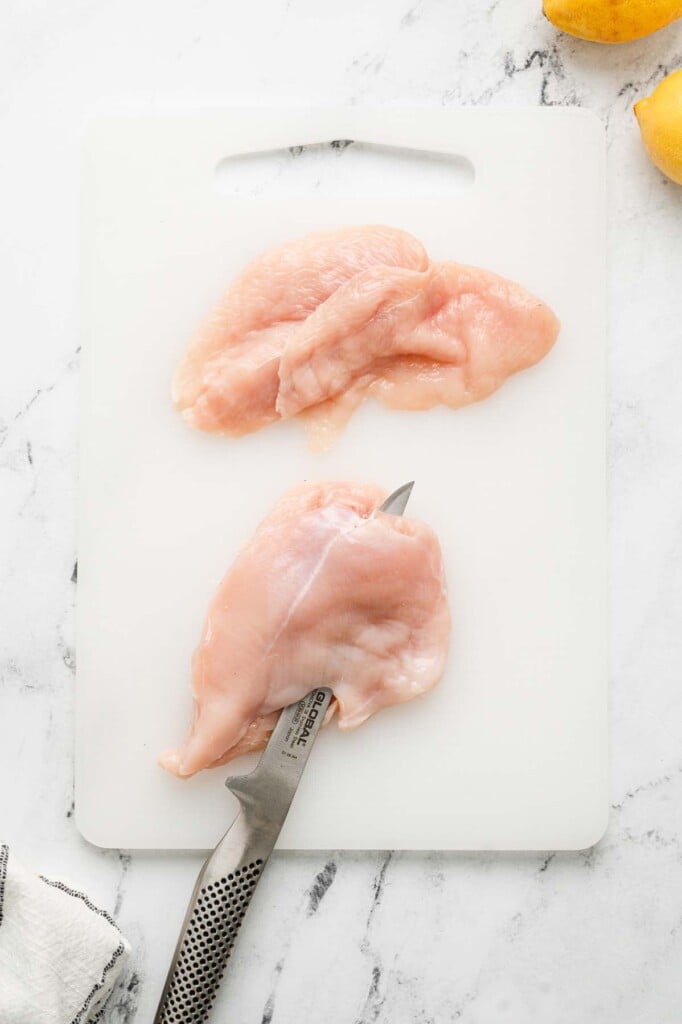 Two chicken breasts on a white cutting board. One is being filleted 