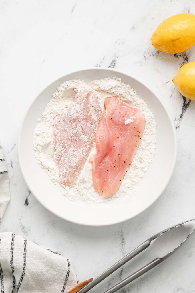 Two chicken breast getting dredged in flour in a white shallow bowl with lemons on the side