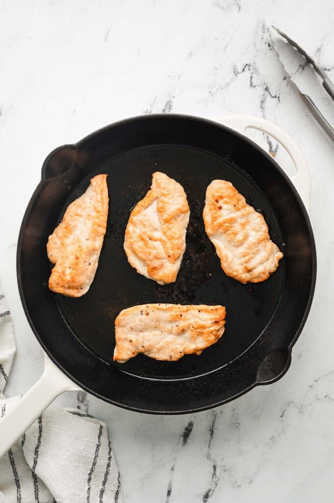 four chicken breasts getting browned in a cast iron pan on a white backgorund