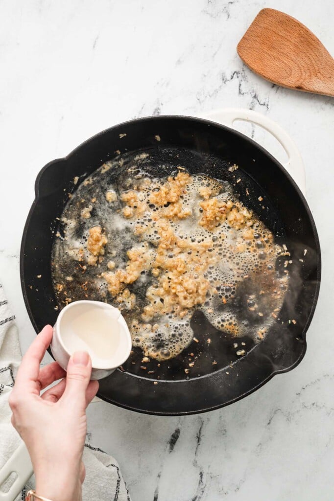 shallots and garlic getting cooked in a cast iron skillet with a hand pouring white wine in it to make chicken piccata sauce