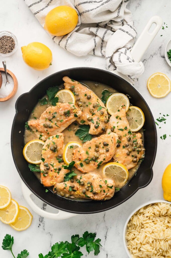 An overhead shot of a cast iron pan of chicken piccata with lemon and cooked orzo on the side