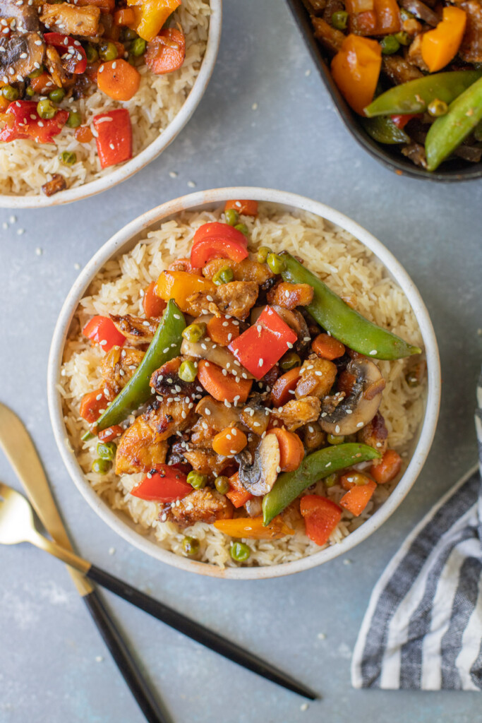 Chicken Stir Fry on a bed of rice in a bowl with another bowl and skillet on the side on a grey background