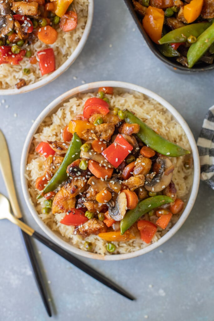Chicken Stir Fry on a bed of rice in a bowl with another bowl and skillet on the side on a grey background