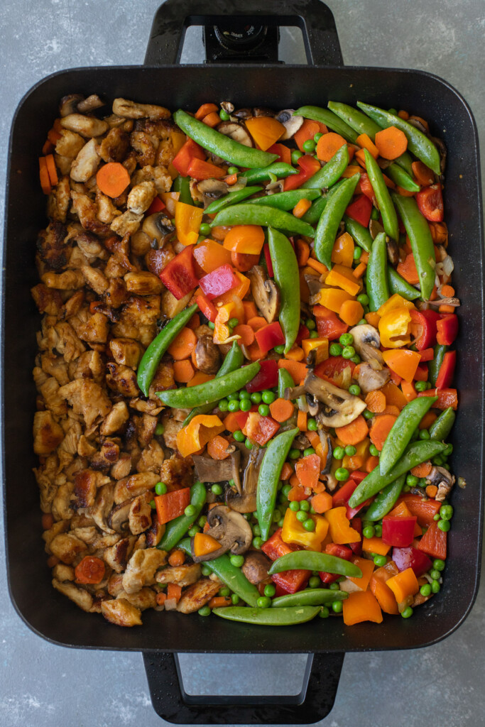 Chicken on half an electric frying pan with a variety of vegetables on the other half getting stir fried