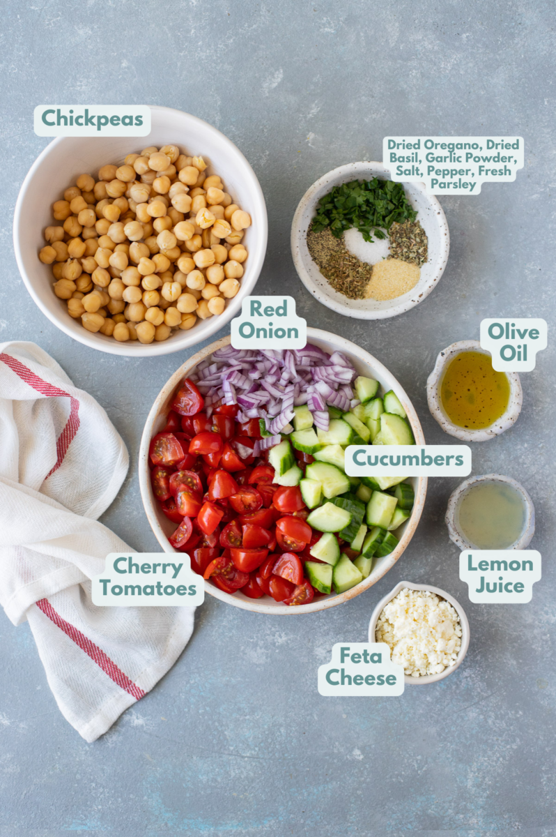 Ingredients for Mediterranean chickpea salad in small bowls: red onion, cherry tomatoes, cucumbers, chickpeas, olive oil, lemon juice, garlic powder, dried basil, dried oregano, salt, pepper, fresh parsley, crumbled feta