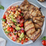 An overhead shot of a plate with grilled chicken thighs on half and greek chickpea salad on the other half on a grey background