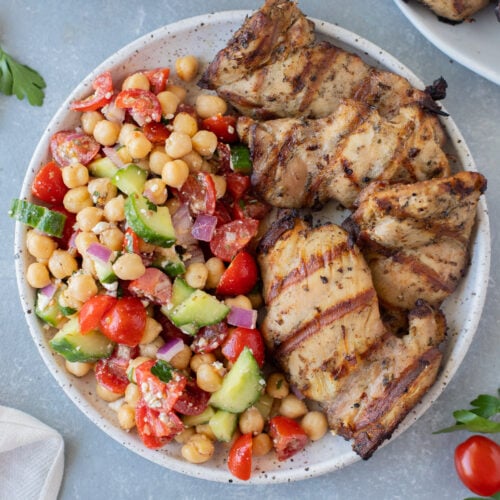 An overhead shot of a plate with grilled chicken thighs on half and greek chickpea salad on the other half on a grey background