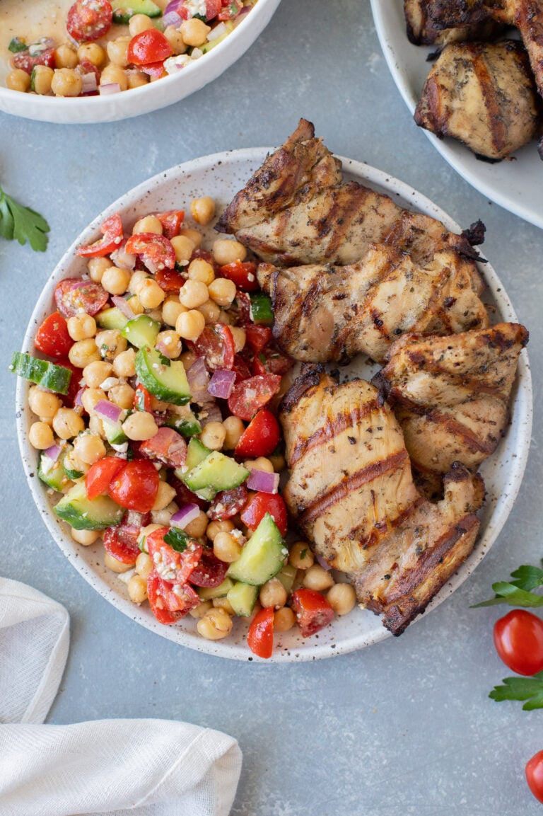 An overhead shot of a plate with grilled chicken thighs on half and greek chickpea salad on the other half on a grey background