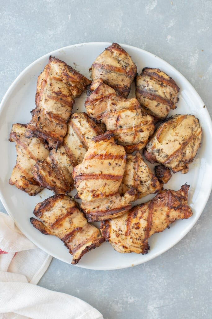 An overhead shot of a white plate of grilled boneless chicken thighs on a grey background