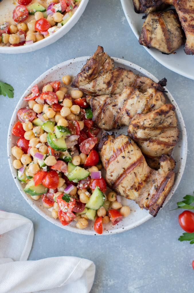 An overhead shot of a plate with grilled chicken thighs on half and greek chickpea salad on the other half on a grey background with a plate of the chicken on the side