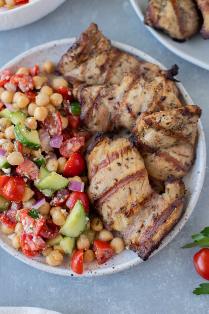A close up of a plate with grilled chicken thighs on half with Mediterranean chickpea salad on the other half on a grey background