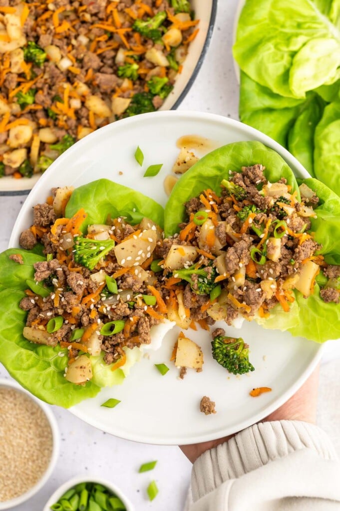 Two Ground Beef Lettuce Wraps on a white plate being held up by a hand with the pan of wrap filling in the background