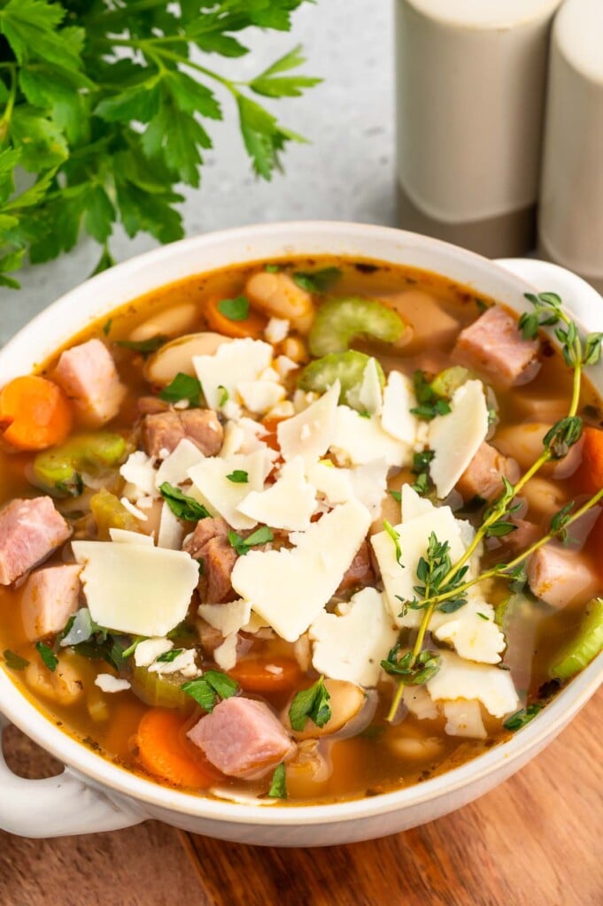 A white bowl of Ham white bean soup on a wooden cutting board with salt and pepper shakers and fresh parsley in the background