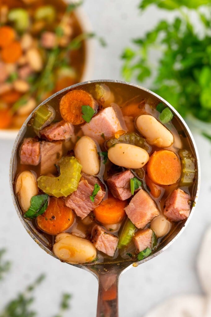 A ladle holding up a scoop of ham white bean soup with another bowl of the soup in the background