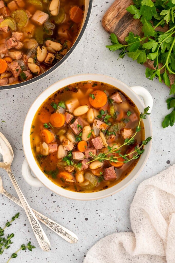 A white bowl of ham white bean soup with the pot of soup on the side on a white counter