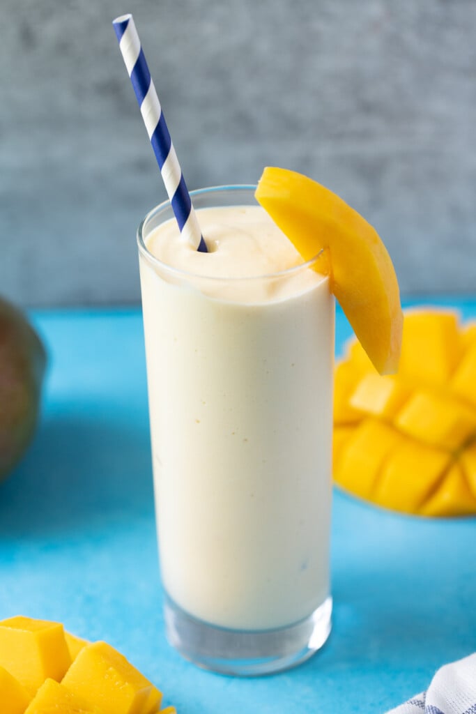 A mango smoothie on a blue background with mangos off to the side and a blue paper straw