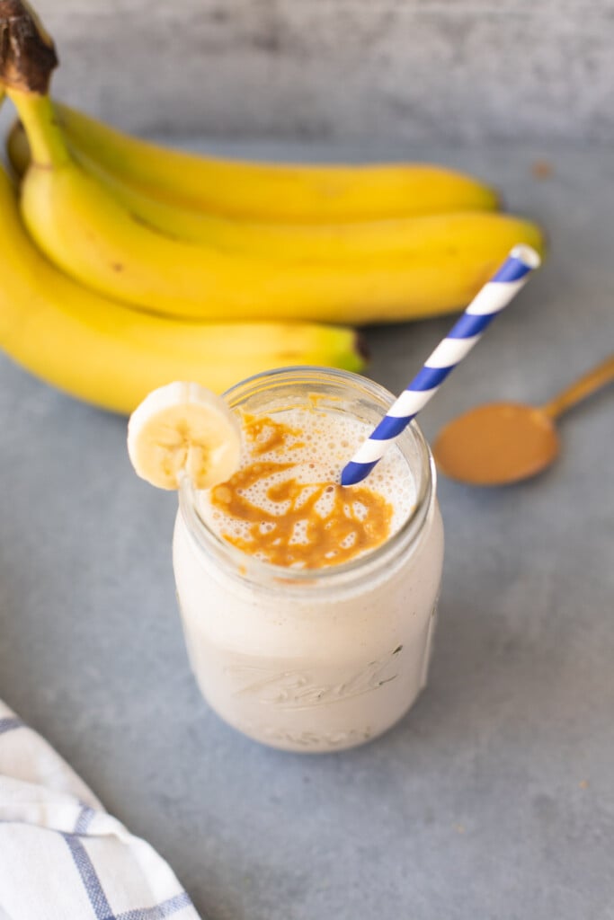 A banana smoothie in a mason jar with a blue striped straw and bananas in the background