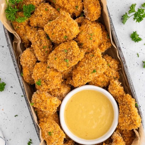An overhead shot of a platter of chicken nuggets with a white bowl of honey mustard on a white background