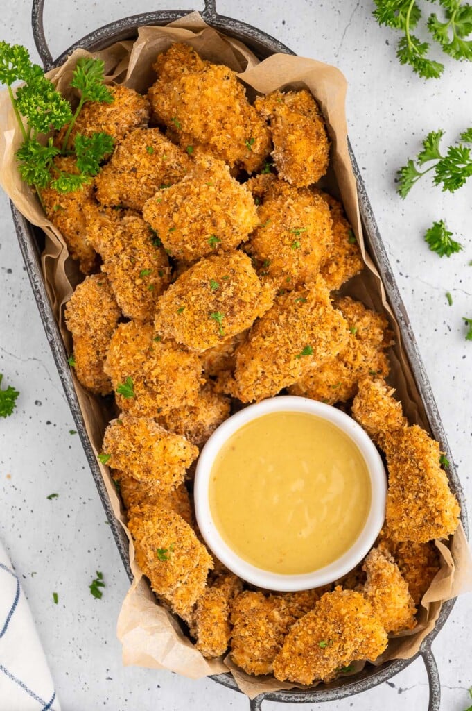 An overhead shot of a platter of chicken nuggets with a white bowl of honey mustard on a white background