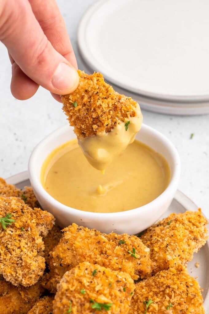 A hand dipping a homemade healthy chicken nugget into a white bowl of honey mustard with more chicken nuggets on the side on a white background