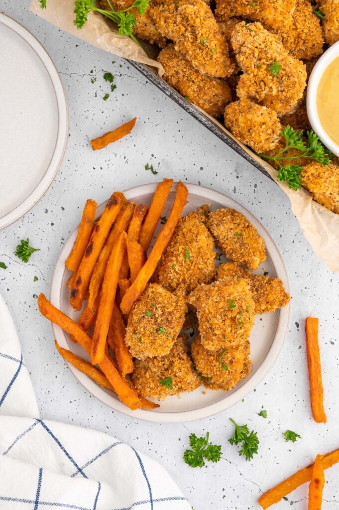 An overhead shot of a white plate with chicken nuggets and sweet potato fried with a platter of chicken nuggets on the side on a white background