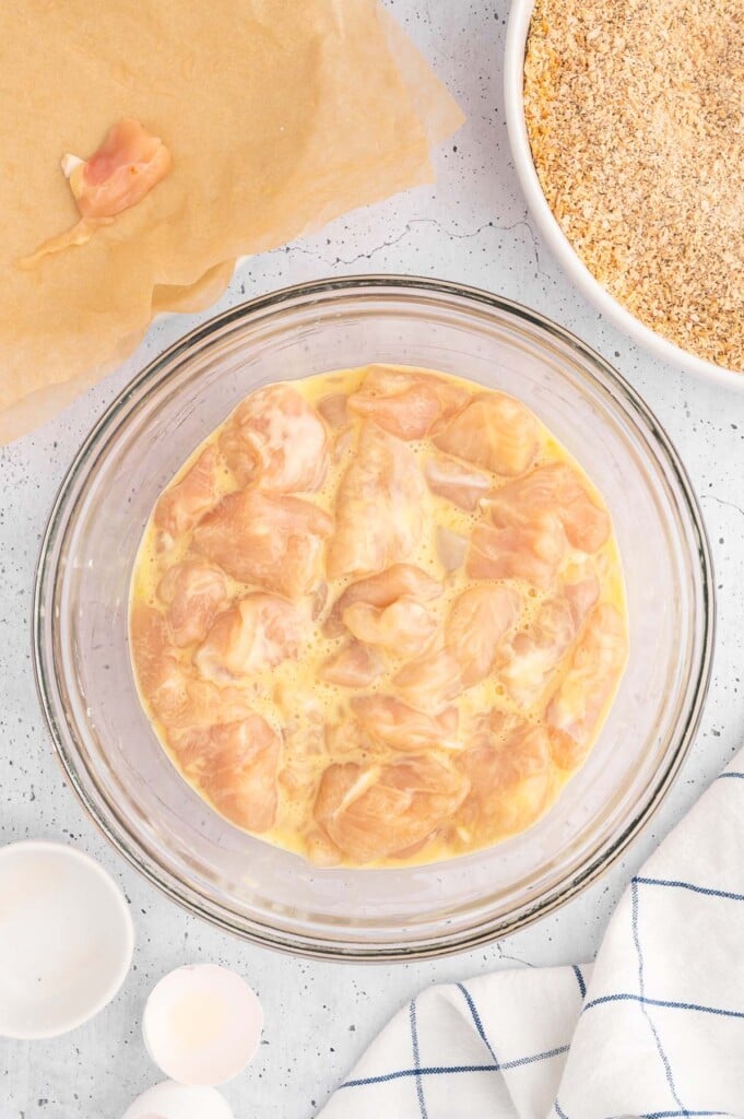 Raw chicken tenderloins cut into small inch nuggets in a clear bowl with egg and milk with starch sprinkled over the top to make chicken nuggets