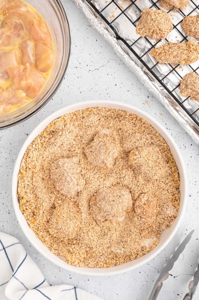 Homemade chicken nuggets getting dipped in a breadcrumb mixture before baking with a wire raking on a baking sheet on the side