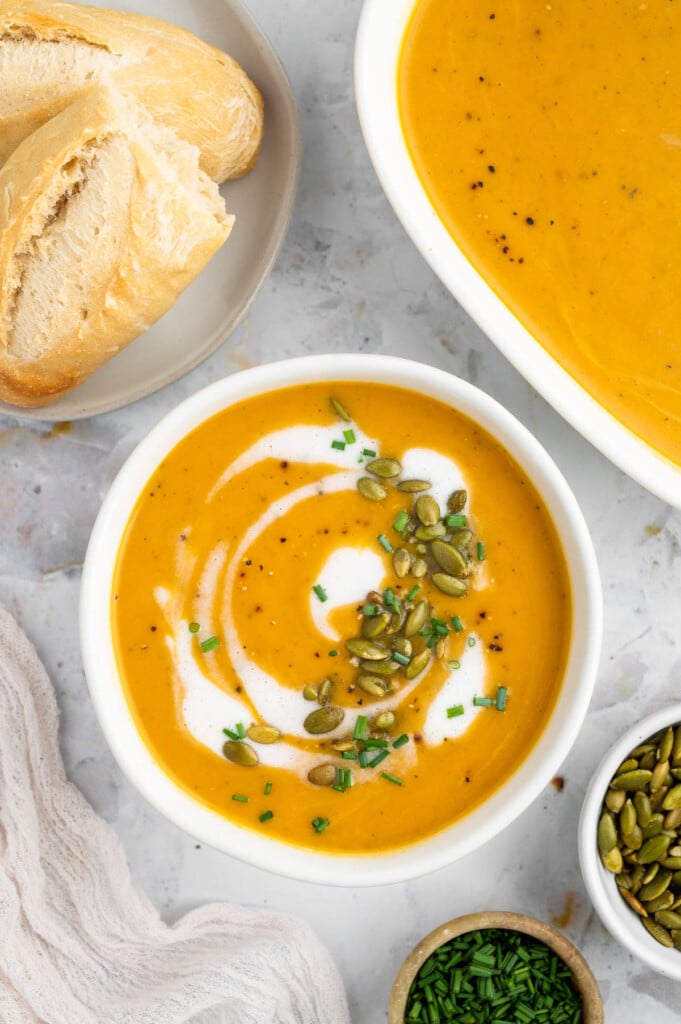 An overhead shot of a white bowl of instant pot butternut squash soup with cream, herbs and pumpkin seeds on top with a dish of soup and bread on the side for one pot meals